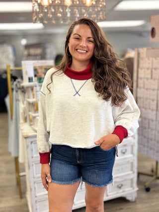 A woman modeling an ash grey colored knit crewneck sweater with contrasting dark red ribbed collar, cuffs, and hem, paired with blue denim shorts in a boutique setting.
