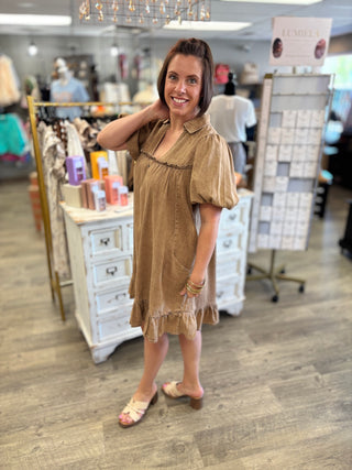 A woman modeling a light brown short-sleeve dress with puff sleeves and a ruffled hem inside a boutique store.
