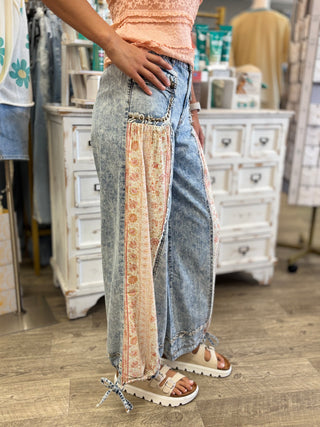 Model wearing light-wash denim patchwork pants with floral patterned fabric panels and frayed details, paired with beige slide sandals in a boutique setting.