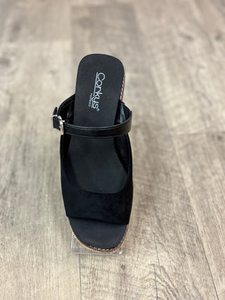 A black wedge sandal with a slingback strap, displayed on a clear stand over a gray wood-look floor.