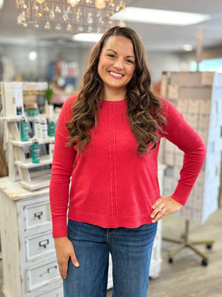 A smiling woman with long wavy brown hair stands in a boutique wearing a bright red long-sleeve knit top and blue jeans.