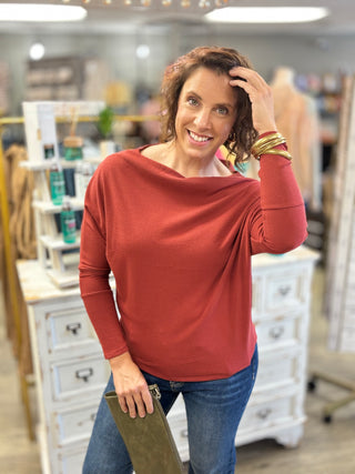 A woman wearing a rust-orange long-sleeve top and blue jeans stands in a retail store holding a brown clutch. The top has a wide draped neckline and is the primary product shown.