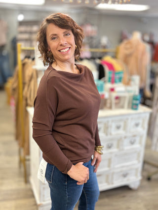 A woman with short brown hair smiling in a clothing boutique, wearing a brown long-sleeve top and blue jeans.