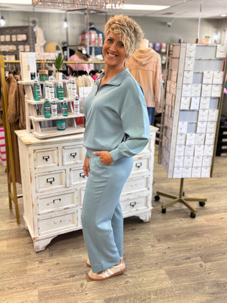 A woman modeling a light blue matching lounge set consisting of a long-sleeve collared top and wide-leg pants, standing inside a retail store.