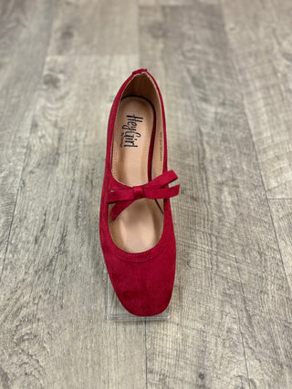 A single red suede Mary Jane flat shoe with a matching bow detail across the instep, displayed on a clear acrylic block over a gray wood-look floor. 