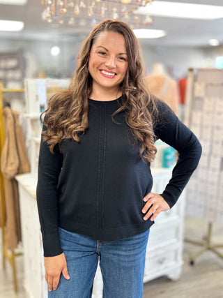 A woman with long wavy brown hair smiling in a boutique, wearing a black long-sleeve crewneck sweater and blue jeans.