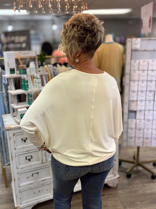 A woman viewed from behind wearing a cream-colored, dolman top and blue jeans inside a retail store.