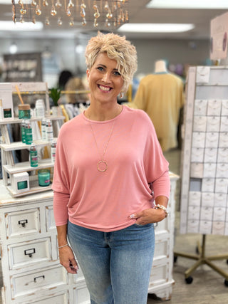 A smiling woman with short blonde hair stands in a boutique store wearing a dusty pink long-sleeve top and blue jeans. She is wearing a gold circular pendant necklace.
