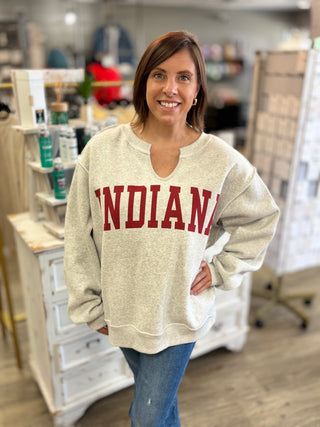 A woman wearing a light heather gray sweatshirt with 'INDIANA' printed in red across the chest, standing in a retail store.