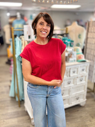 Woman wearing a red shirt and blue jeans standing in a store with clothing racks and mannequins in the background.