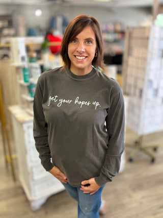 A woman with shoulder-length brown hair stands in a shop wearing a dark charcoal gray long-sleeve crewneck sweatshirt with the phrase 'get your hopes up.' printed in white script across the chest, paired with blue jeans.