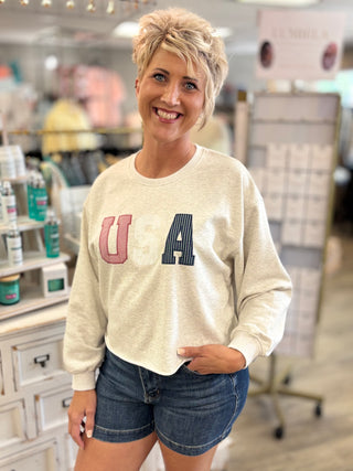 Woman wearing a cream sweatshirt with 'USA’ in red, white, and blue letters in a store setting.