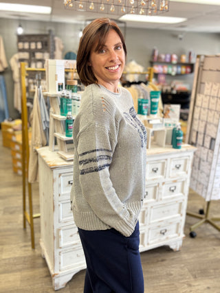A woman with short brown hair smiling in a boutique, wearing a light gray knit sweater with navy blue stripes on the sleeves and dark navy pants.