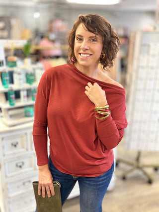 A woman wearing a rust-red off-the-shoulder long-sleeve top and blue jeans, standing in a retail store environment.