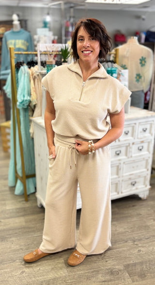 A woman modeling a beige two-piece lounge set consisting of a short-sleeve half-zip top and matching wide-leg pants, standing inside a clothing boutique.
