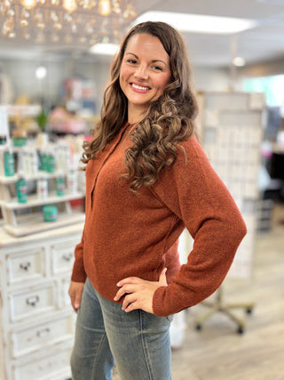 A smiling woman with long wavy brown hair stands in a retail store wearing a rust-orange long-sleeve knit sweater and light blue jeans.