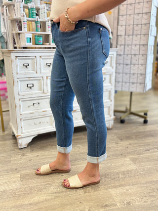 A person wearing medium-wash blue cuffed jeans and cream slide sandals, standing in a retail store with a white cabinet and wood floor visible.