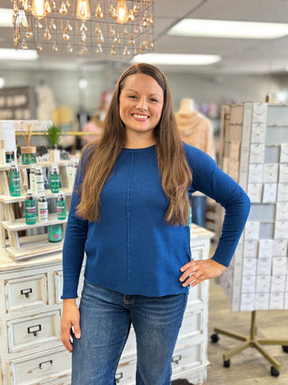 A woman with long light brown hair stands in a retail store wearing a royal blue long-sleeve knit top and blue jeans. The background shows store fixtures, display shelves with products, and a hanging light fixture.