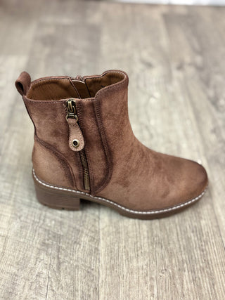 A brown suede ankle boot with a side zipper and pull tab, shown from a side profile on a light wood floor.