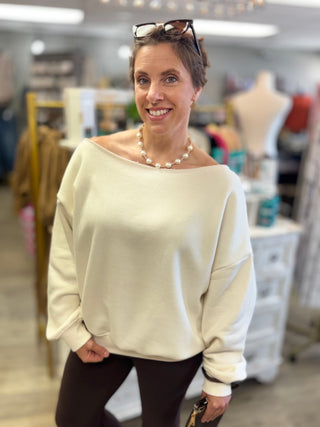 A woman smiling in a clothing boutique, wearing a cream off-the-shoulder knit sweater, brown pants, and a pearl necklace.