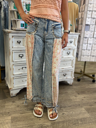 Model wearing wide-leg patchwork jeans with a light blue denim base and beige floral vertical panels, paired with a peach top and sandals in a clothing store.