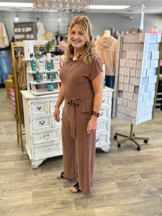 A woman modeling a brown short-sleeve jumpsuit with a tie waist, standing in a retail store setting with white display cabinets and clothing racks in the background.