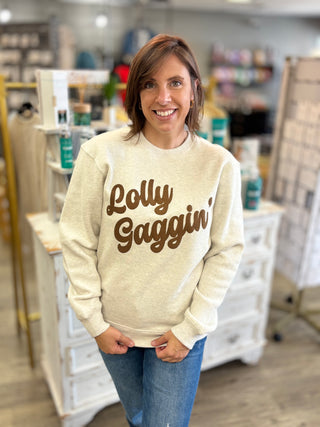 A woman smiling in a boutique while wearing a cream-colored crewneck sweatshirt with 'Lolly Gaggin' printed in brown script across the chest, paired with blue jeans.