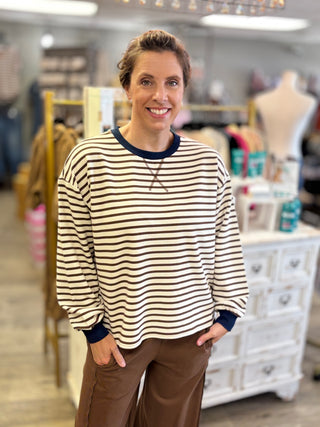A woman smiling in a boutique while wearing a cream and brown horizontally striped long-sleeve shirt with navy trim details and brown pants.
