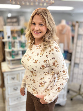 A smiling blonde woman wearing a cream-colored long-sleeve knit sweater with a brown and tan floral pattern, standing in a boutique setting.