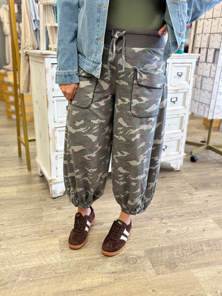 A person wearing loose-fit camouflage cargo jogger pants with a drawstring waist, styled with a denim jacket, olive green shirt, and brown sneakers in a boutique setting.