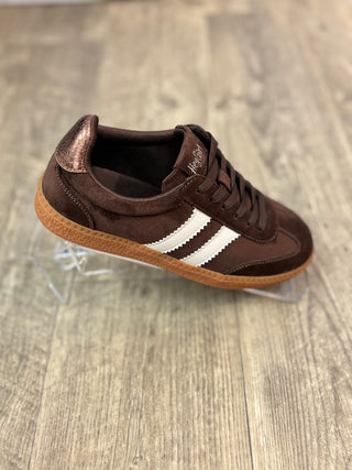 A dark brown suede sneaker with three white stripes on the side, resting on a clear plastic display stand over a gray wood-look floor.