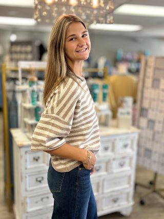 A woman with blonde hair smiling and looking over her shoulder, wearing a beige and white striped knit top and dark blue jeans, standing inside a boutique shop.