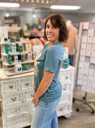 A woman with short brown hair smiling and looking over her shoulder in a retail store, wearing a teal graphic t-shirt and light blue jeans. Shelves with products are visible in the background.