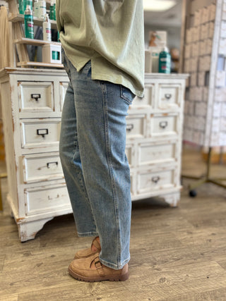 A person wearing light-wash blue straight-leg jeans, a light sage green top, and tan suede loafers, standing in a retail store with white wooden cabinets in the background.