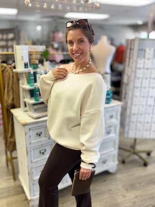 A woman wearing an off-the-shoulder cream knit sweater and dark brown pants, standing in a boutique store.