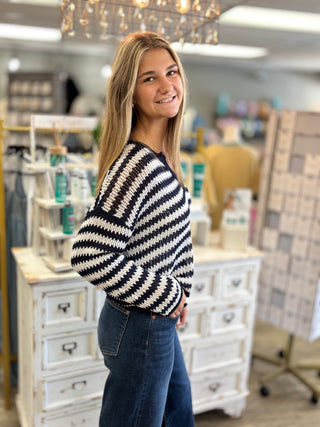 A woman with blonde hair stands in a boutique shop wearing a black and white striped knit sweater and dark blue jeans.