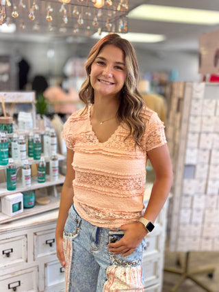 A woman smiling in a boutique store, wearing a peach-colored ruffled lace short-sleeve top and light-wash jeans.