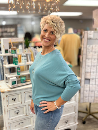 A smiling woman with short blonde hair stands in a retail store wearing a teal top and blue jeans, posing next to a white wooden display cabinet.