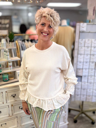 A woman with short blonde hair smiling in a boutique, wearing a cream-colored textured knit top with ruffle details and striped pants.
