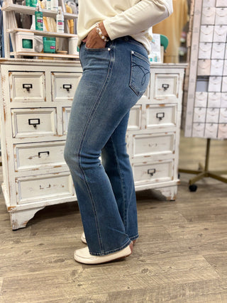 A person wearing medium-wash blue flare jeans, standing in a shop with a white vintage-style drawer cabinet behind them.