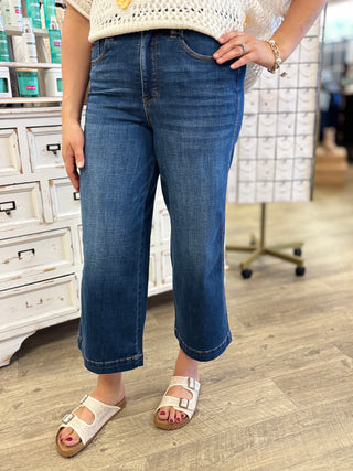 A person wearing dark blue cropped denim jeans with a straight-to-wide leg fit, paired with a cream knit top and white slide sandals in a retail store setting.