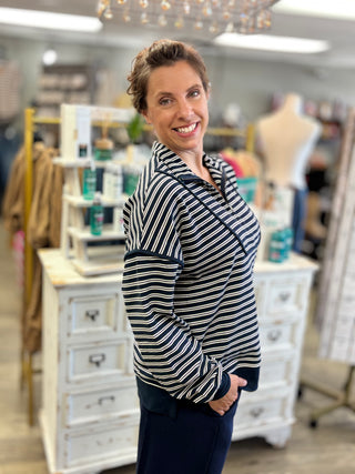 A woman smiling and posing in a boutique, wearing a navy and white vertically striped zip-up top and dark navy pants.