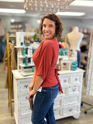 A woman with short brown hair smiling and looking back over her shoulder in a boutique. She is wearing a rust-red draped top with a side tie and blue jeans, standing in front of a white wooden cabinet.