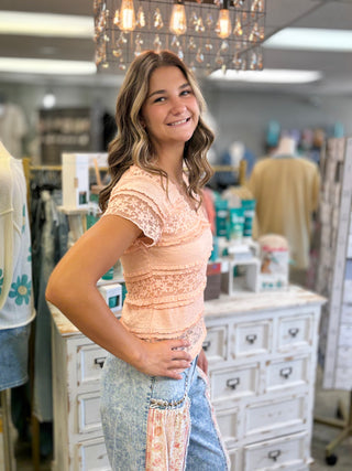 A woman posing in a boutique shop, wearing a peach lace short-sleeve top and light blue jeans with a floral patterned side panel.