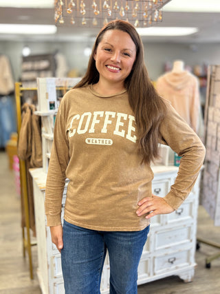 A woman with long brown hair stands in a retail store wearing a tan long-sleeve graphic shirt that says 'COFFEE WEATHER' in cream lettering, paired with blue jeans.