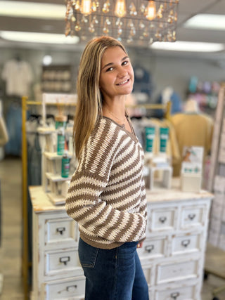 A young woman with long blonde hair smiling and looking over her shoulder, wearing a brown and cream striped knit sweater and blue jeans inside a retail store.