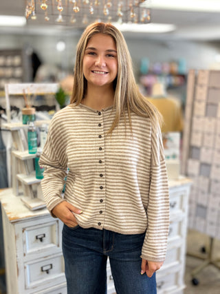 A smiling young woman with blonde hair stands in a boutique shop wearing a beige and white striped long-sleeve button-up cardigan with dark buttons, paired with dark blue jeans.