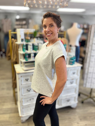 A woman modeling a light grey short-sleeve top and black leggings inside a boutique store with white furniture and display racks in the background.