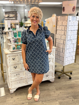 A woman modeling a short-sleeve navy blue dress with a white geometric trellis pattern, standing in a boutique store.