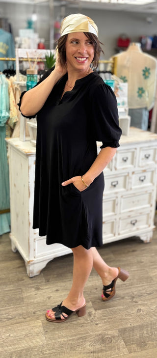 A woman modeling a black short-sleeve knee-length dress with a V-neck and side pockets in a boutique setting. She is wearing a gold patterned headscarf and black slide sandals.